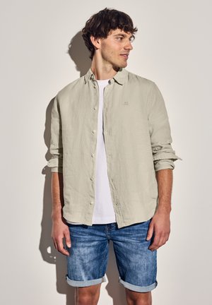 Camisa de botón de manga larga en un tono gris claro con mangas enrolladas, combinada con una camiseta blanca y pantalones cortos de mezclilla. Diseño sencillo, ajuste casual.