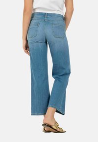 Lichtblauwe wijde jeans met een middelhoge taille, met achterzakken en subtiele stiksels, gedragen met bruine platte sandalen.