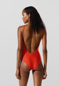Rød badedragt i ét stykke med tynde stropper, højt skåret ben og dyb åbning bagpå. Glat tekstur med minimalistisk design.