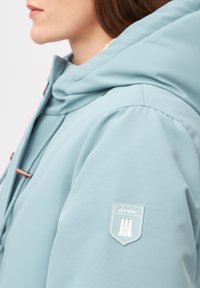 Ljusblå hoodiejacket med en slät yta, prydd med en dekorerande patch på vänster ärm och bronserad hårdvara för dragskojustering.