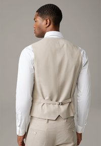 Beige maßgefertigte Weste mit strukturierter Oberfläche, die ein doppelt geknöpftes Design und einen verstellbaren Rückengurt aufweist. Weiße Hemdsärmel sind sichtbar.