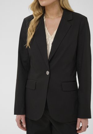 Femme portant un blazer noir à un bouton sur un haut clair à sequins avec les cheveux blonds lâchés, vue des épaules aux hanches.