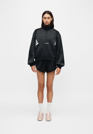 Kvinde står med front mod kameraet iført sort Adidas træningsjakke og shorts, hvide sokker og lyserøde sneakers foran en ensfarvet lys baggrund.
