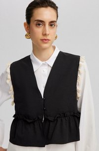 Chaleco negro con fondo volante y acentos de borlas en los hombros, usado sobre una camisa blanca de cuello. Presenta cierres de botones en la parte frontal.