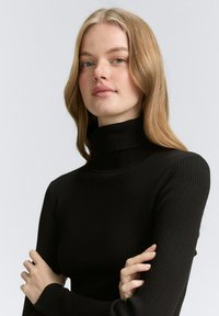 Pull à col roulé noir côtelé à manches longues, avec un col haut et une coupe ajustée. La texture est lisse et extensible.