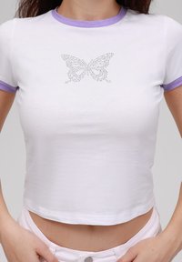 Camiseta blanca cropped con cuello de canalé morado, que presenta un diseño de mariposa hecho de strass en el frente. Tejido de algodón suave.