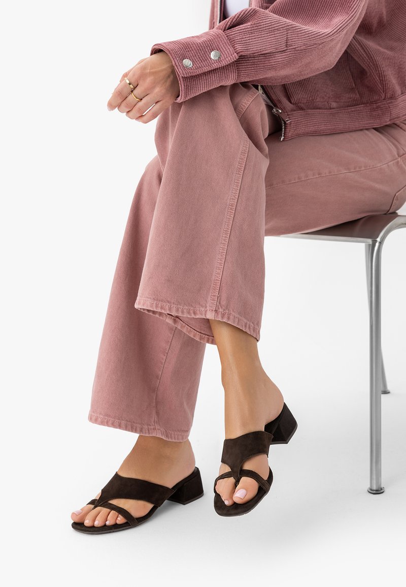 Femme assise sur une chaise en métal, portant un pantalon large rose, une veste en velours côtelé rose et des sandales à talons ouvertes noires avec de larges brides.