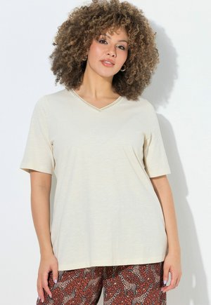 Vrouw met krullend haar, gekleed in een beige T-shirt met korte mouwen en V-hals en een broek met giraffenpatroon, staande tegen een witte achtergrond.