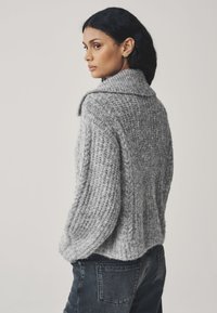 Maglione grigio lavorato a maglia con ampio colletto, trama a coste e orlo leggermente corto, indossato con jeans scuri.