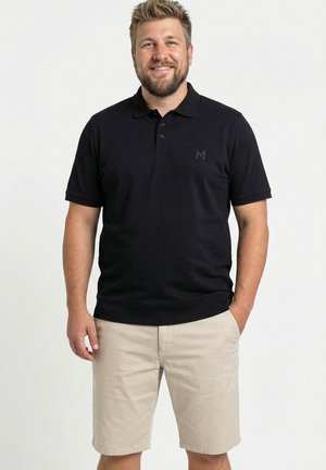 Mand med skæg, smilende, iført sort poloshirt med korte ærmer og beige shorts, stående mod en hvid baggrund.