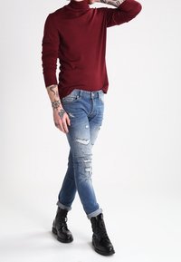 Maglione dolcevita borgogna, aderente, abbinato a jeans chiari strappati. Stivali neri con lacci, orli arrotolati, tatuaggi visibili sull'avambraccio.