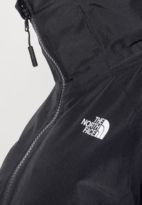 Veste imperméable noire avec une fermeture éclair à l'avant et une capuche. Le logo The North Face est affiché en blanc sur la partie gauche de la poitrine.