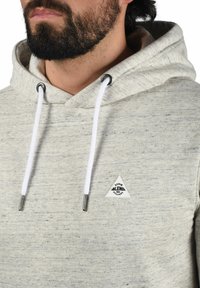 Lichtgrijze hoodie met een textuurafwerking, voorzien van een driehoekig logo-embleem en witte trekkoorden met metalen uiteinden.