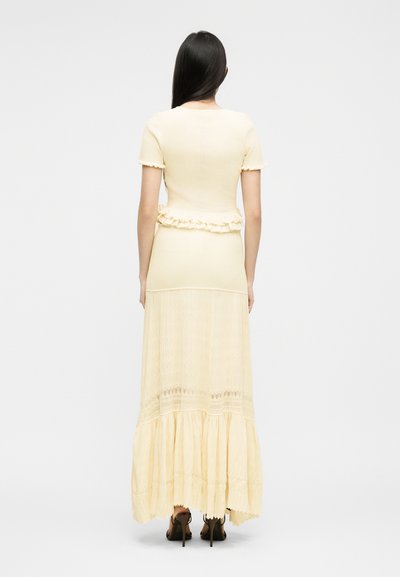 Γυναίκα με μακριά σκουρόχρωμα μαλλιά φοράει ανοιχτό κίτρινο φόρεμα με κοντά μανίκια, λεπτομέρεια με βολάν στη μέση και μακριά φούστα, σε συνδυασμό με μαύρες ψηλοτάκουνες γόβες.