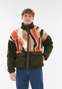 Chaqueta de forro polar con un patrón abstracto multicolor en naranja, verde y beige. Cuenta con bolsillos con cremallera y un diseño de cuello alto.