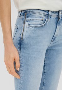Gros plan d'une personne portant un jean en denim bleu clair avec une poche zippée verticale sur le côté avant gauche et une main reposant à côté.