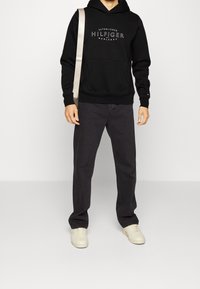 Felpa nera con logo bianco "Hilfiger", tasca frontale, abbinata a pantaloni grigio scuro e sneakers color beige chiaro. Il look è informale.