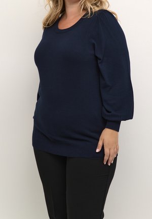 Kvinde iført navyblå langærmet sweater med puffede ærmer og sorte bukser, stående mod en ensfarvet lys baggrund.