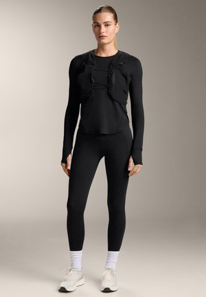 Femme aux cheveux tirés en arrière portant un haut noir à manches longues, un gilet ajusté avec poches avant, des leggings noirs, des chaussettes blanches et des baskets blanches.