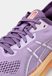 Zapatilla deportiva morada con una parte superior de malla, tela texturizada, acentos blancos y una suela de goma con una entresuela naranja. Los cordones son trenzados.