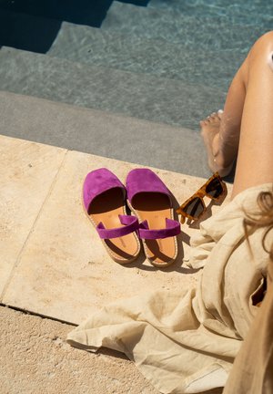 Des chaussures plates en daim violet avec une bride, une semelle plate et une semelle intérieure en cuir beige, posées sur une surface en pierre près d'une piscine. Une paire de lunettes de soleil en écaille de tortue à proximité.