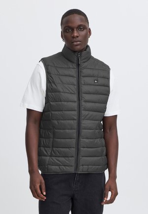 Homme portant une veste noire matelassée sans manches sur un t-shirt blanc et un pantalon noir, debout devant un fond clair uni.