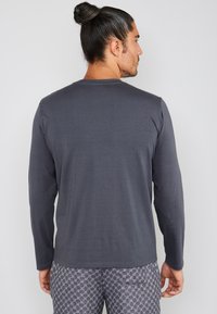 Chemise grise à manches longues avec un col rond. Tissu doux, design minimaliste. Portée avec un pantalon de pyjama à motifs, visible au niveau de la taille.