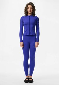 Ljubičasta sportska odjeća koja se sastoji od dugih rukava s patentnim zatvaračem i visoko pojasnim tajicama. Glatka, prilagođena tkanina i minimalistički šavovi. Crne cipele koje se lako obuvaju.