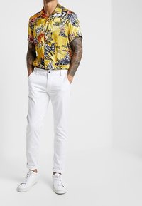 Koszulka z krótkim rękawem w żółte kwiaty z czarnymi i różowymi akcentami, połączona z białymi spodniami slim-fit i białymi trampkami.