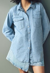 Abito camicia in denim blu chiaro con maniche lunghe, chiusura con bottoni a scatto, due tasche sul petto e orlo curvo. Tessuto testurizzato con finitura morbida.