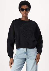 Melna sporta krekls ar teksturētu apļa logotipu priekšā, ar ribu aprocēm un apakšmalu; apvienots ar gaiši zilām augsta vidukļa džinsām.