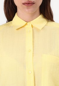 Camicia gialla con bottoni, caratterizzata da un colletto classico, tessuto testurizzato, taschino frontale e un aspetto leggero e trasparente.