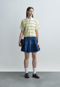 Mladá žena v žlto pruhovanom polo tričku, modrej džínovej sukni na gombíky, bielych ponožkách, čiernych mokasínach, drží čiernu kabelku na sivom pozadí.