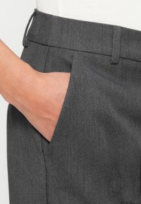 Main insérée dans la poche avant d’un pantalon gris foncé sur mesure avec passants de ceinture visibles et détails de couture.