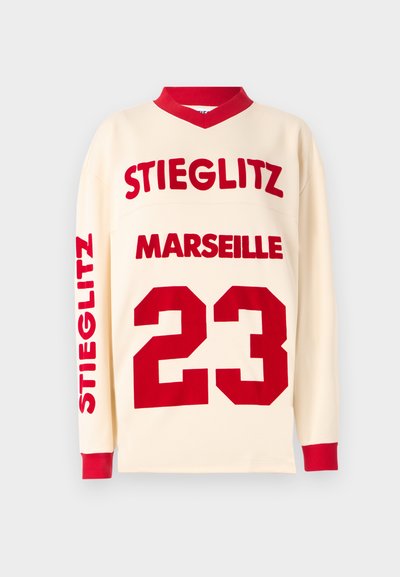 Krémová košeľa s dlhým rukávom a červenými akcentmi. Obsahuje veľký červený text "STIEGLITZ" a "MARSEILLE", pričom číslo "23" je výrazne zobrazené.