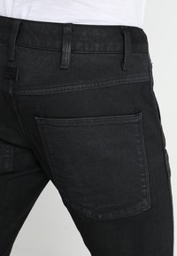 Mustat farkut, joissa on teksturoitu pinta, yksittäinen takatasku ja vyölenkit, esittelevät tyylikästä, kapeaa leikkausta.