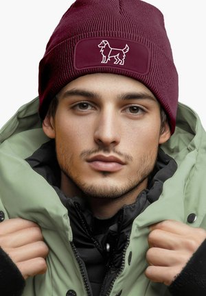 Burgunderfarbene Strickmütze mit Hundemotiv, getragen von einer Person in einer grünen Jacke. Die Mütze hat eine gerippte Struktur und ein flaches Patch.