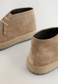 Bottines en suède brun clair avec une surface texturée, dotées de lacets à l'avant et d'une semelle en caoutchouc naturel avec des accents beiges.