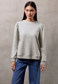 Pullover grigio con un texture a pois, scollo tondo e maniche a sbuffo. Abbinato a jeans a gamba larga blu su uno sfondo neutro.
