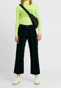 Personne portant un haut à manches longues vert fluo, un pantalon à carreaux vert foncé et noir, des baskets vert fluo et un sac bandoulière noir, se tenant contre un fond blanc.