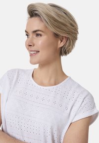 Weißes Baumwolloberteil mit aus den Stoff ausgeschnittenen Bestickungen und rundem Halsausschnitt. Kurze Ärmel mit umgeschlagenem Saum und strukturiertem Muster.