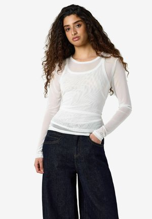 Jeune femme aux longs cheveux bouclés portant un haut blanc transparent à manches longues par-dessus un débardeur blanc et un jean large foncé, debout avec une main dans la poche.