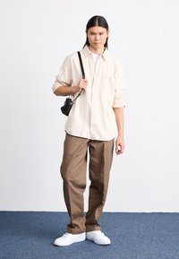 Beige gestreiftes Hemd mit Knöpfen, lockere braune Hose und weiße Sneakers. Kleine schwarze Umhängetasche mit Kettenriemen. Einfarbiger Hintergrund.