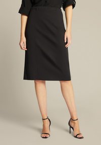 Schwarzer knielanger Bleistiftrock aus glattem Stoff, mit einer figurbetonten Silhouette und elastischem Bund. Dazu werden schwarze Riemchen-High Heels getragen.