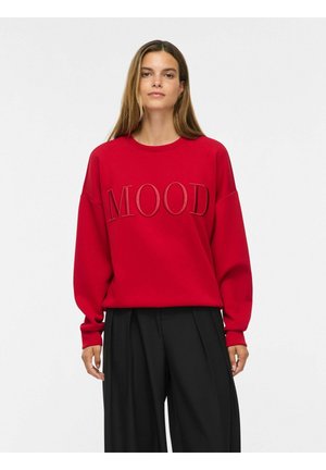 Femme aux longs cheveux bruns portant un sweat-shirt rouge avec "MOOD" brodé et un pantalon noir à jambes larges, debout devant un fond uni.
