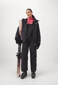 Schwarze Skijacke über schwarzen Skihosen, mit einer pink gemusterten Innenlage. Hält Skier, trägt schwarze Skischuhe und Handschuhe.