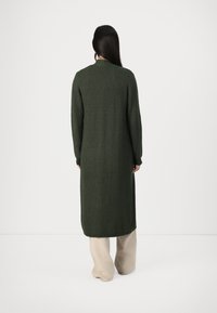 Lang, ribbet cardigan i mørkegrønn, med høy hals og lange ermer, kombinert med beige, vide bukser.