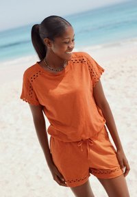 Orange bomuldsoutfit med en kortærmet top med bølgemønster og matchende shorts med udskæringer, sat mod en strandbaggrund.