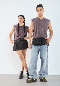 To modeller står sammen. Kvinnen har på seg en lilla ermeløs topp, svarte denimskjørt og svarte støvler. Mannen har på seg en lignende topp sammen med baggy blå jeans og flerfargede sneakers. Begge har tilbehør som belte og en svart hofteveske.