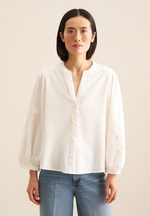 Weiße Bluse mit Knopfleiste, rundem Ausschnitt und puffärmeligen Ärmel mit bestickten Details an den Bündchen. Getragen zu blauen Jeans.